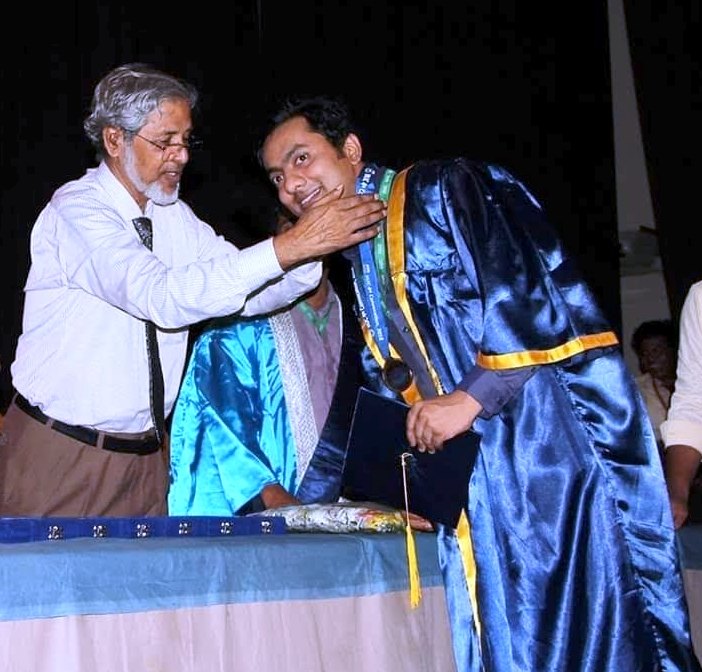 At the convocation ceremony: a university officer places the medal ribbon over the shoulders of the graduand, who wears an academic gown and holds his certificate.